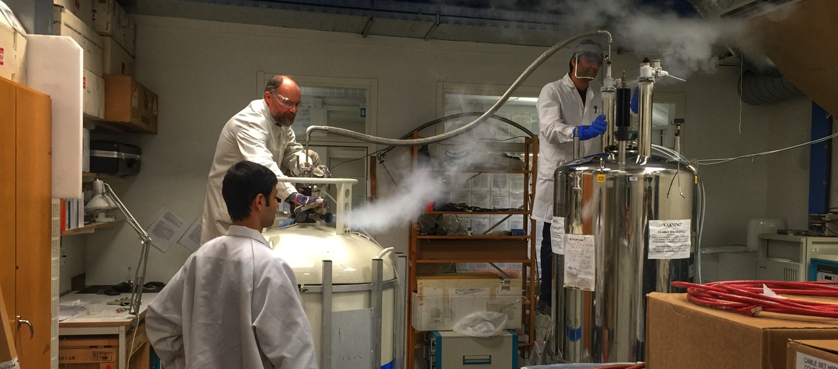 tre person i vita rockar och skyddsglasögon i ett labb som håller på och gör tester. Vit rök kommer ut från ena tuben.