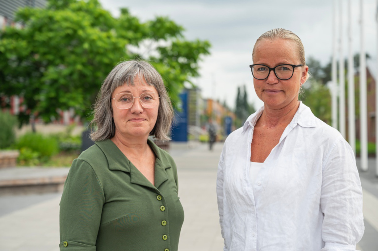 Kristina Ek och Linda Wårell står utomhus och tittar in i kameran och ler, Luleå campus i bakgrunden, träd och gångbana syns bakom