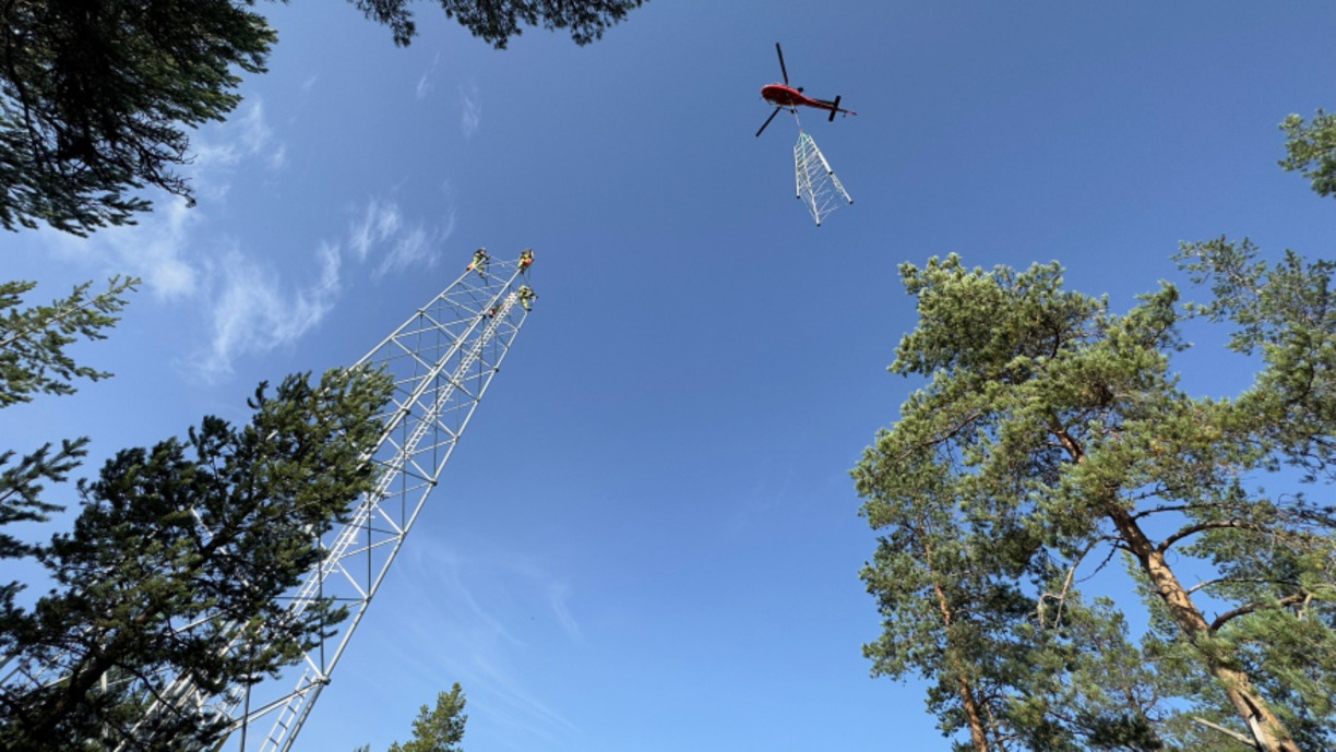 Helikopter med en mast hängandes under åker ner mot marken. En mast står på plats intill träd.