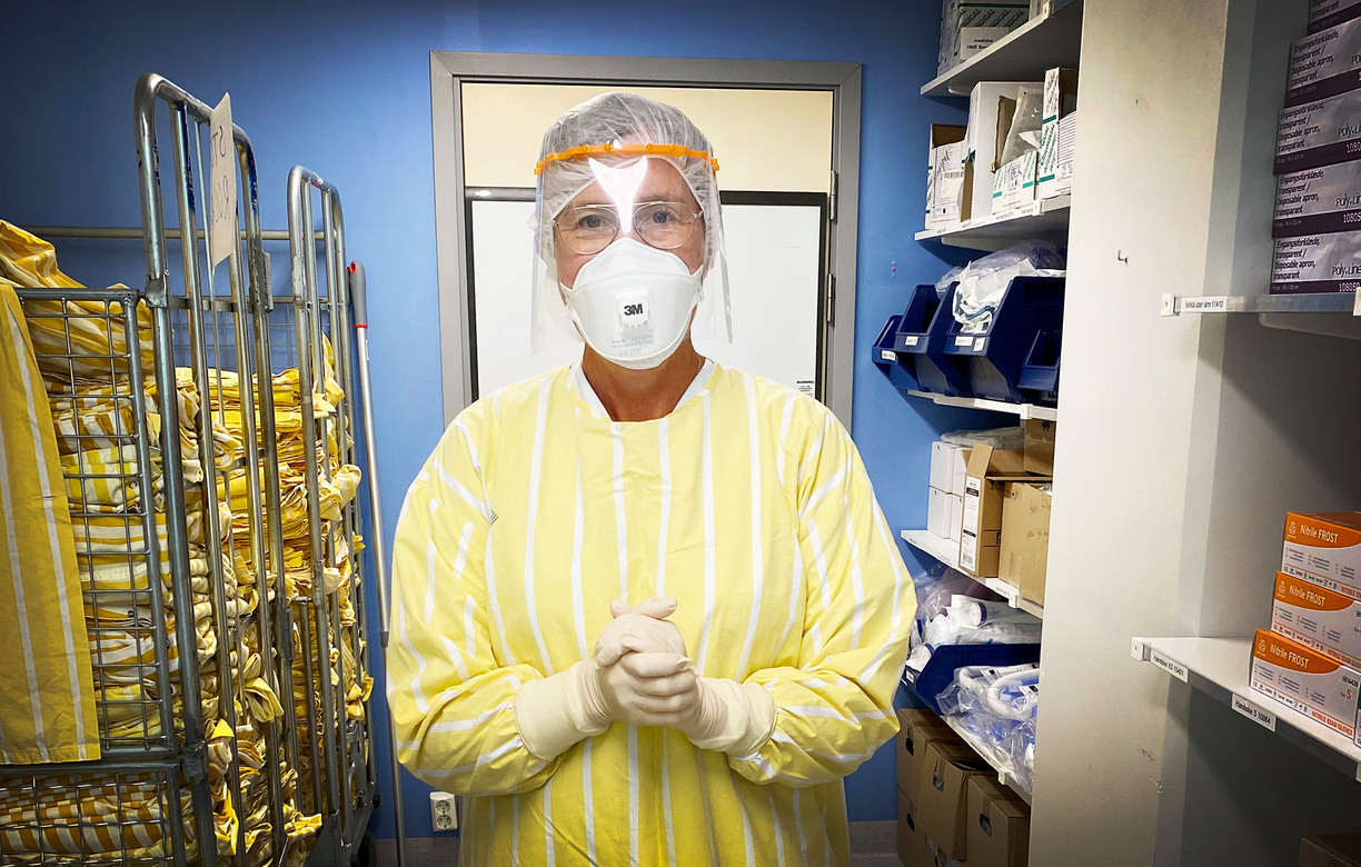 Kvinna stående med skyddskläder och mask för ansiktet under Corona pandemin, står i ett mindre rum med fler rockar och utrustning