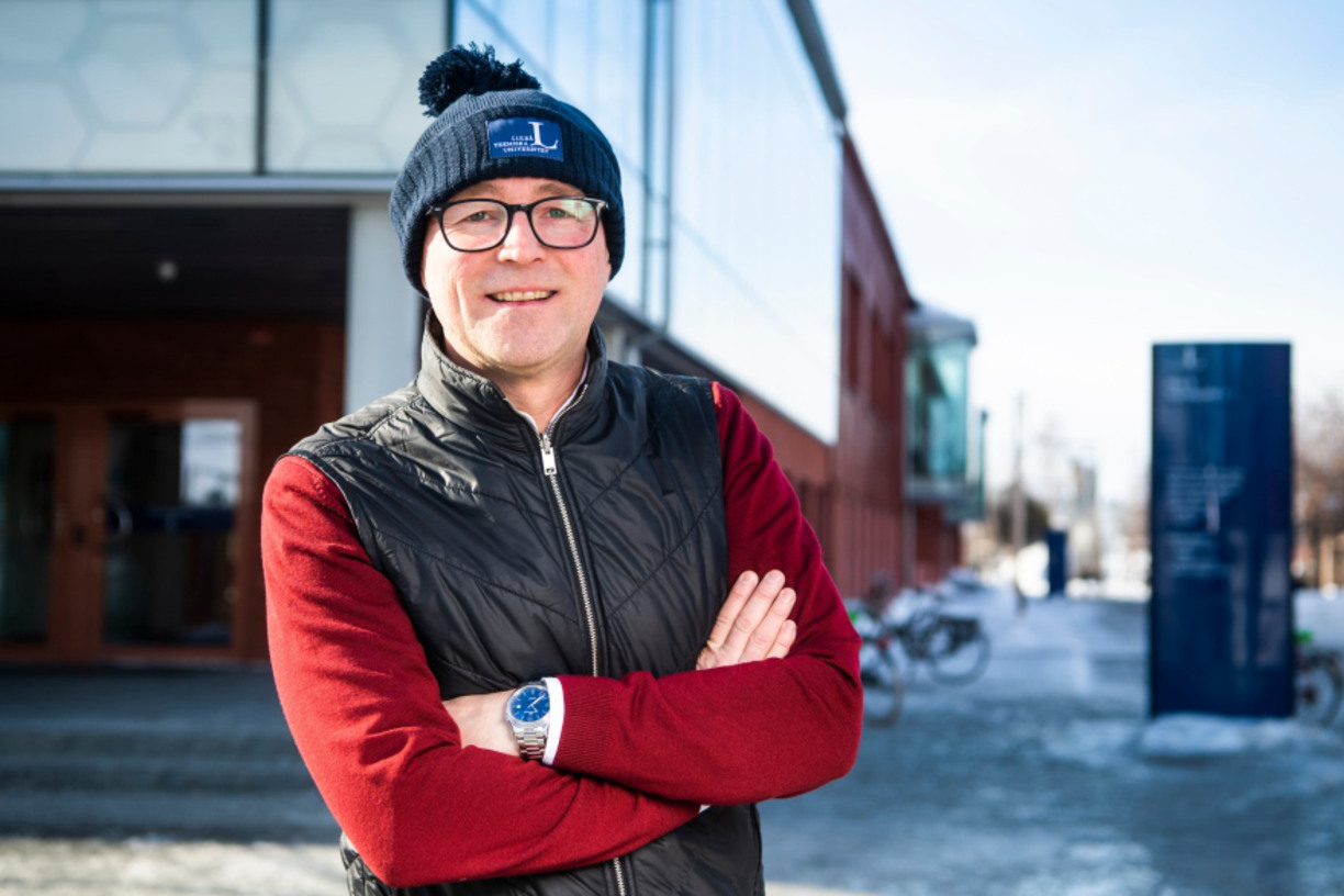 Joakim Abrahamsson smiles into the camera, with the LTU logo on his hat, in the background a building and some snow