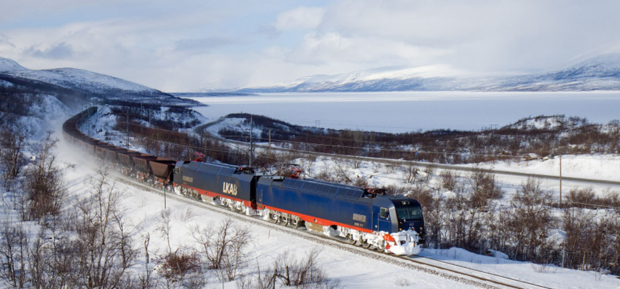Översiktsbild på ett tåg som åker i ett vintrigt landskap. Berg syns i bakgrunden