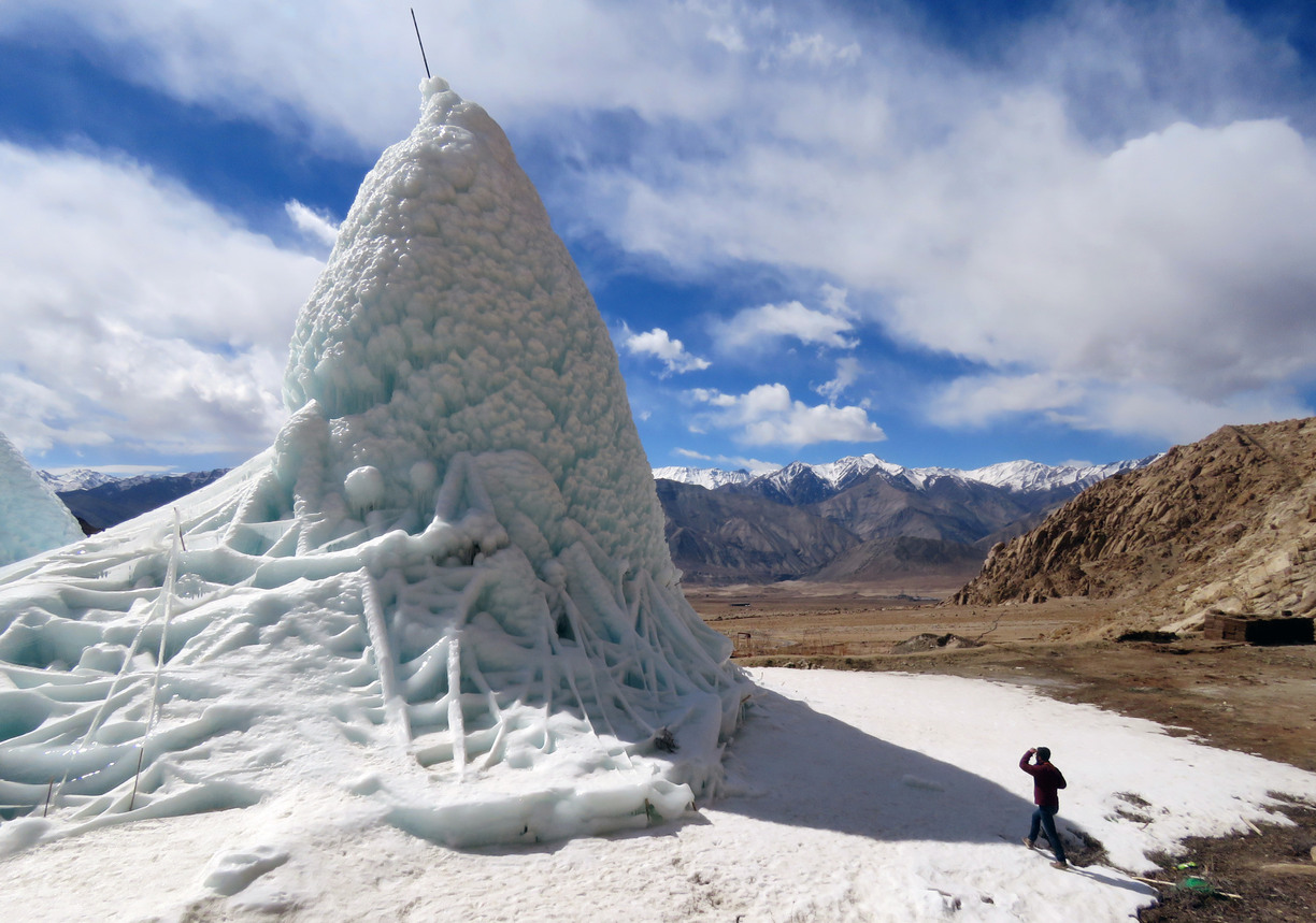 Glaciär främst i bild, bakom syns berg och en person som står nedanför glaciären