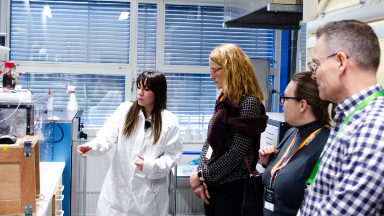 Io Antonopoulou står i labb i labbrock och visar tre personer som står och lyssnar
