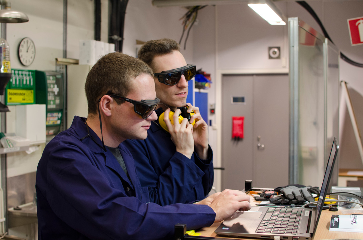Två forskare iklädda labbrock och skyddsutrustning, står vid en dator i ett laboratorie